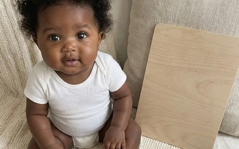A baby in a white bodysuit sitting on a cream knitted blanket beside a blank wooden milestone board and dried flowers